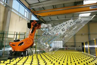 Orange robot arm with a suction frame beside a large glass panel and yellow rollers.