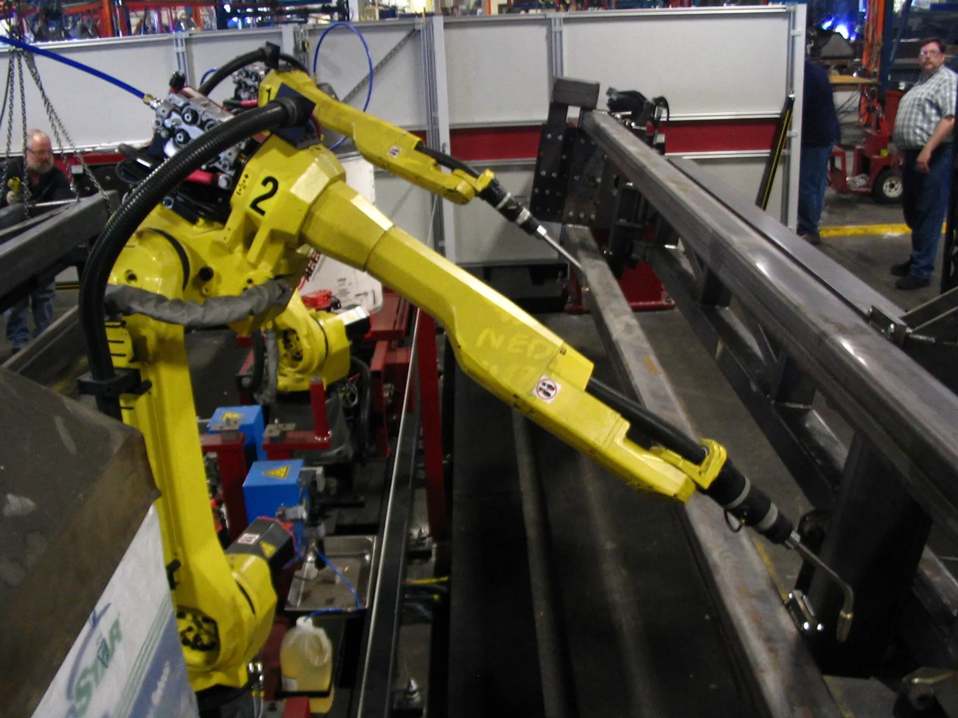 Yellow robot arms beside a long metal structure, with people in the background.