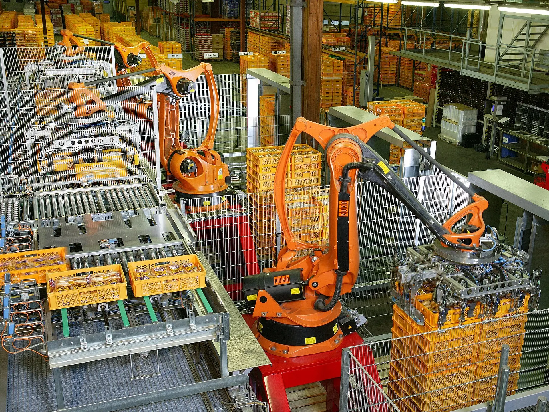 Orange robot arms beside stacked yellow crates and roller conveyors.